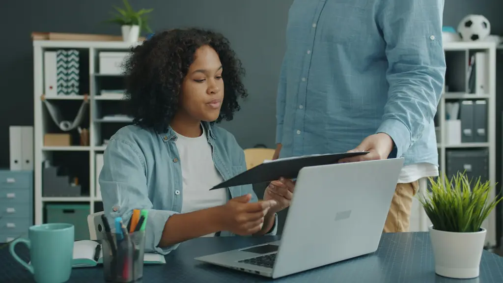 Colleague handing documents to a co-worker at her desk with a laptop
