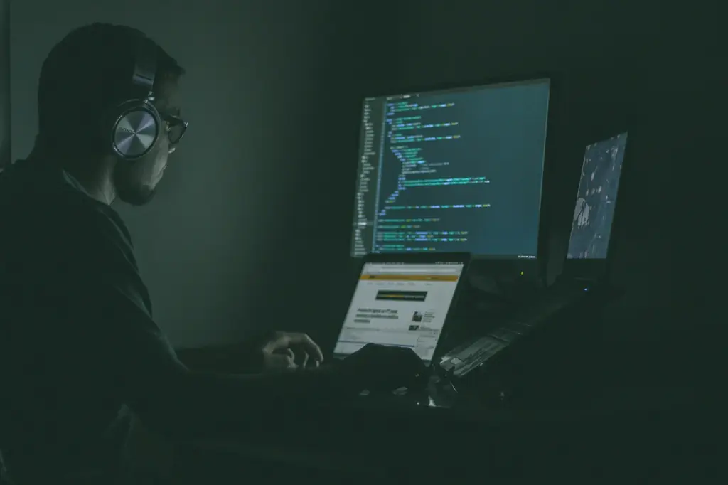 Security tester wearing headphones working at monitors of code in a dark room